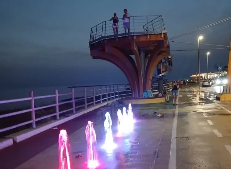 La Skyview di Scoglitti di notte: due persone sulla piattaforma panoramica, davanti giochi d'acqua colorati sul lungomare.