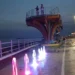 La Skyview di Scoglitti di notte: due persone sulla piattaforma panoramica, davanti giochi d'acqua colorati sul lungomare.