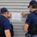 Due agenti di polizia in giubbotto antiproiettile applicano un cartello di chiusura su una serranda metallica.