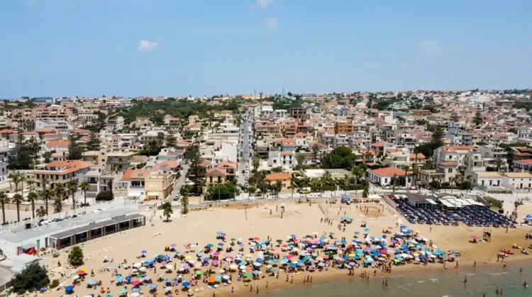 Vista aerea della spiaggia di Marina di Ragusa con bagnanti, ombrelloni colorati e il centro abitato sullo sfondo.