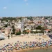Vista aerea della spiaggia di Marina di Ragusa con bagnanti, ombrelloni colorati e il centro abitato sullo sfondo.