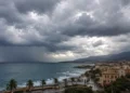 Nuvole temporalesche su un centro costiero siciliano con mare mosso, cortina di pioggia all'orizzonte e palme piegate dal vento.