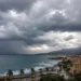 Nuvole temporalesche su un centro costiero siciliano con mare mosso, cortina di pioggia all'orizzonte e palme piegate dal vento.