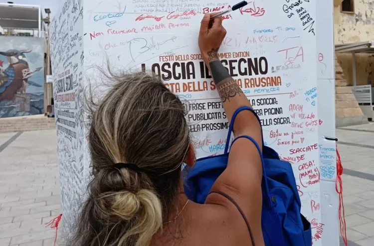 Una turista firma e lascia un messaggio su un totem ricoperto di scritte davanti all'Info Point di Marina di Ragusa.