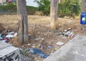 Rifiuti abbandonati tra gli alberi in un'area verde della Villa Margherita di Ragusa, con erba secca e cestini per la raccolta differenziata sullo sfondo