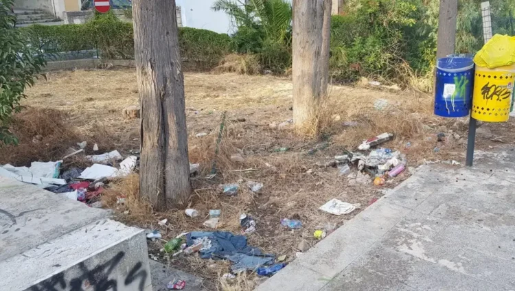 Rifiuti abbandonati tra gli alberi in un'area verde della Villa Margherita di Ragusa, con erba secca e cestini per la raccolta differenziata sullo sfondo