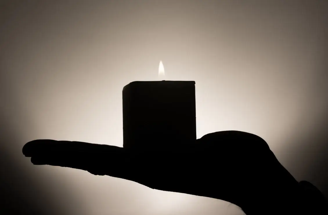 Sagoma di una mano che regge una candela accesa davanti a uno sfondo scuro