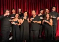 Il cast della Compagnia Godot in abiti scuri, con tazze da tè in mano, davanti a un sipario rosso.
