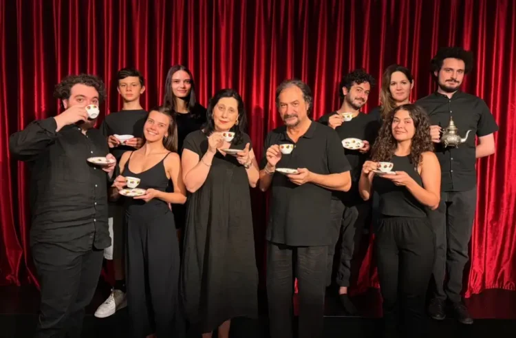 Il cast della Compagnia Godot in abiti scuri, con tazze da tè in mano, davanti a un sipario rosso.