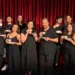 Il cast della Compagnia Godot in abiti scuri, con tazze da tè in mano, davanti a un sipario rosso.