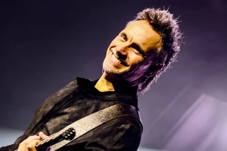 Un cantante sorride sul palco, con il microfono in mano e una chitarra a tracolla.