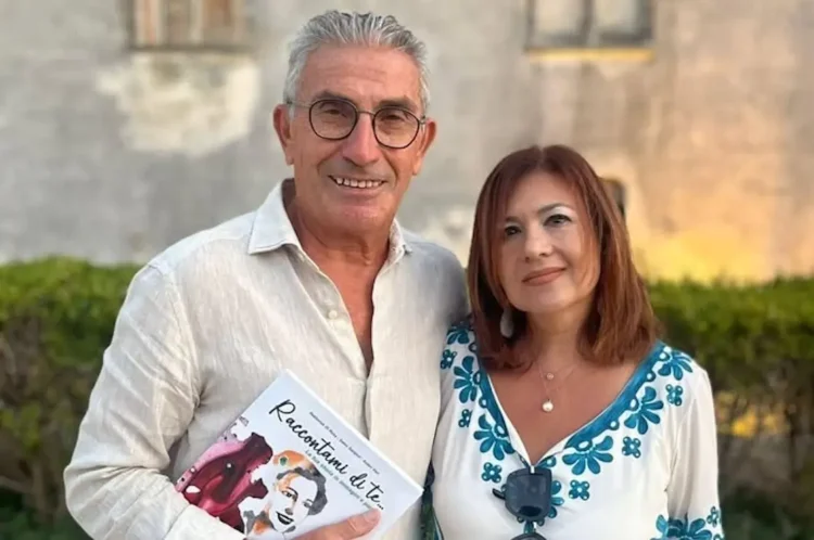 Salvatore Battaglia e Salvatrice Corallo posano insieme tenendo in mano il libro "Raccontami di te" davanti a un edificio in pietra.