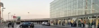 Facciata vetrata del terminal aeroportuale con auto in sosta e persone con bagagli davanti agli ingressi.