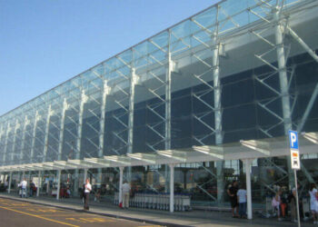 Facciata in vetro e acciaio del terminal dell'aeroporto di Catania Fontanarossa con passeggeri e carrelli portabagagli all'esterno.
