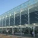 Facciata vetrata del terminal di un aeroporto con struttura in acciaio, viaggiatori e carrelli portabagagli sul piazzale esterno