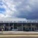 Ingresso dell'Aeroporto Civile di Comiso Pio La Torre con la facciata bianca, alcune auto parcheggiate e cielo nuvoloso