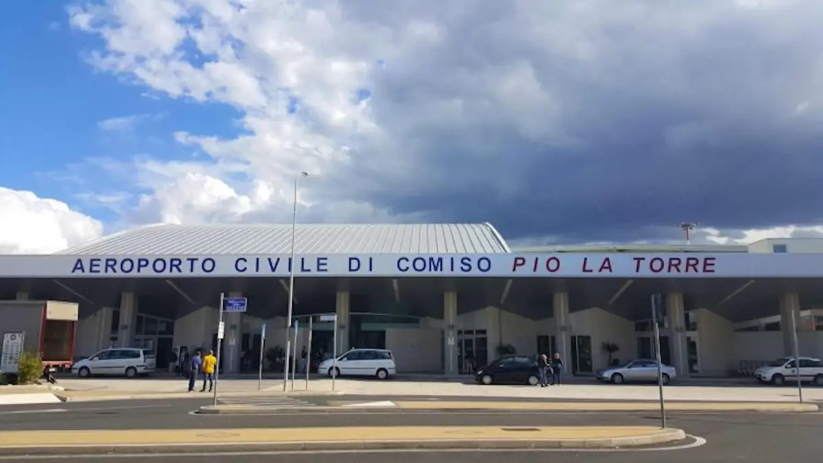 Ingresso dell'Aeroporto Civile di Comiso Pio La Torre con la facciata bianca, alcune auto parcheggiate e cielo nuvoloso
