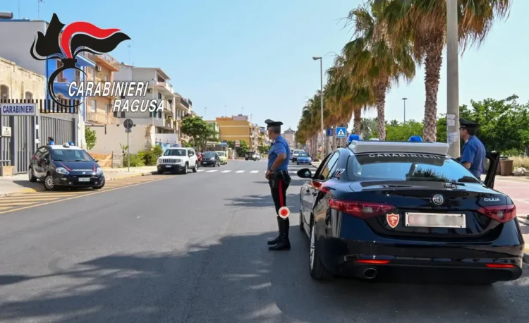 Due auto dei Carabinieri e due militari in uniforme durante un controllo su una strada alberata di Ragusa.