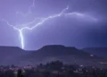 Un fulmine cala su una collina durante un temporale notturno, con un paese illuminato sullo sfondo.