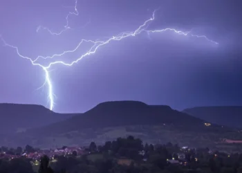Un fulmine cala su una collina durante un temporale notturno, con un paese illuminato sullo sfondo.