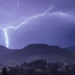Un fulmine cala su una collina durante un temporale notturno, con un paese illuminato sullo sfondo.