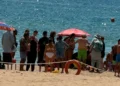 Bagnanti riuniti in cerchio su una spiaggia, salvagente rosso in primo piano, transenna di corda e mare sullo sfondo.