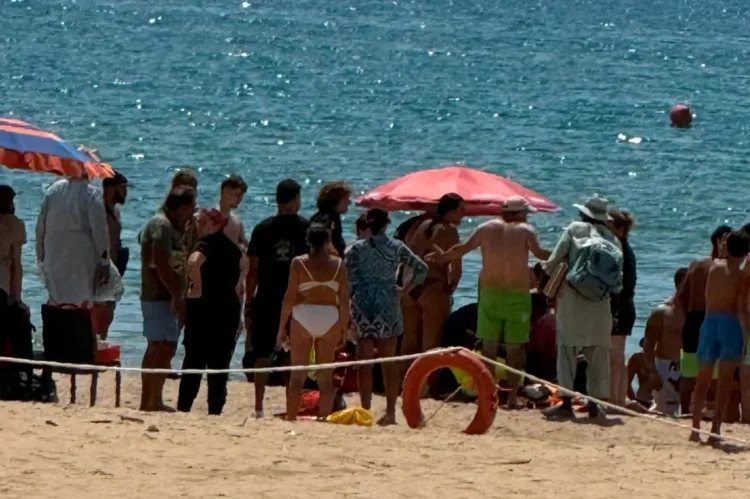 Bagnanti riuniti in cerchio su una spiaggia, salvagente rosso in primo piano, transenna di corda e mare sullo sfondo.