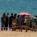 Bagnanti riuniti in cerchio su una spiaggia, salvagente rosso in primo piano, transenna di corda e mare sullo sfondo.