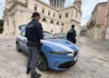 Due agenti della Polizia di Stato accanto a un'auto di pattuglia in una piazza con una chiesa sullo sfondo