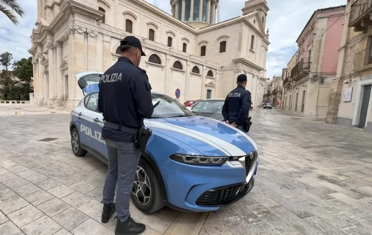 Due agenti della Polizia di Stato accanto a un'auto di pattuglia in una piazza con una chiesa sullo sfondo