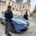 Due agenti della Polizia di Stato accanto a un'auto di pattuglia in una piazza con una chiesa sullo sfondo