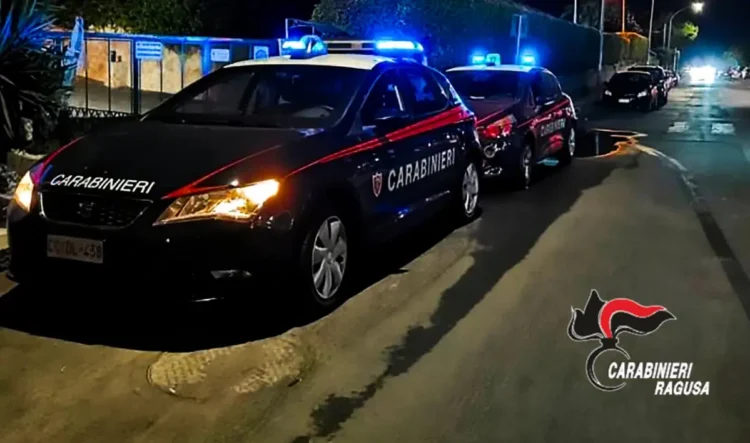 Due auto dei Carabinieri con lampeggianti blu accesi in una strada di notte, logo Carabinieri Ragusa in basso a destra.