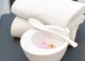 Asciugamani bianchi piegati accanto a una ciotola con miscela rosa, cucchiaio e piccoli fiori.