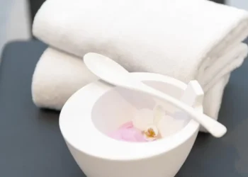 Asciugamani bianchi piegati accanto a una ciotola con miscela rosa, cucchiaio e piccoli fiori.