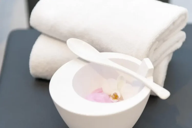 Asciugamani bianchi piegati accanto a una ciotola con miscela rosa, cucchiaio e piccoli fiori.