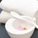 Asciugamani bianchi piegati accanto a una ciotola con miscela rosa, cucchiaio e piccoli fiori.