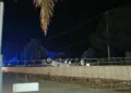 Automobile capovolta di notte dietro un recinto in cemento, persone e mezzi di soccorso sul posto.