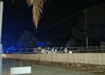 Automobile capovolta di notte dietro un recinto in cemento, persone e mezzi di soccorso sul posto.