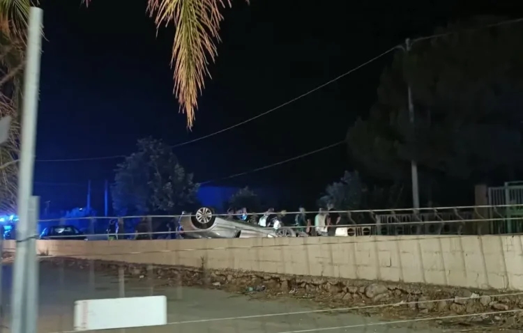 Automobile capovolta di notte dietro un recinto in cemento, persone e mezzi di soccorso sul posto.