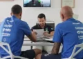 Due atleti e un tecnico seduti a un tavolo esaminano documenti, con uno schermo che mostra un test su cyclette sullo sfondo