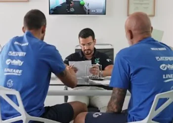 Due atleti e un tecnico seduti a un tavolo esaminano documenti, con uno schermo che mostra un test su cyclette sullo sfondo