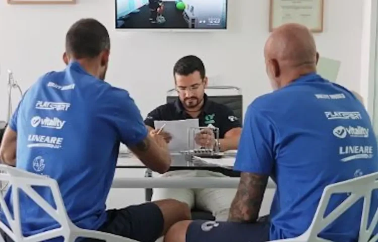 Due atleti e un tecnico seduti a un tavolo esaminano documenti, con uno schermo che mostra un test su cyclette sullo sfondo