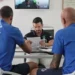 Due atleti e un tecnico seduti a un tavolo esaminano documenti, con uno schermo che mostra un test su cyclette sullo sfondo