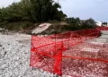 Recinzione arancione da cantiere sulla spiaggia di ciottoli di Playa Grande, con lastre in pietra e vegetazione sullo sfondo.