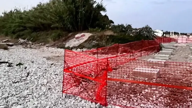 Recinzione arancione da cantiere sulla spiaggia di ciottoli di Playa Grande, con lastre in pietra e vegetazione sullo sfondo.