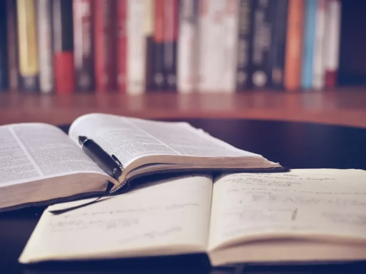 Libri aperti su un tavolo con una penna appoggiata sulle pagine e un quaderno con appunti scritti a mano in primo piano.
