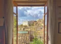 Vista dal centro storico di Modica attraverso una finestra aperta: tetti in cotto, un palazzo bianco e un campanile.