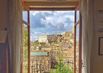Vista dal centro storico di Modica attraverso una finestra aperta: tetti in cotto, un palazzo bianco e un campanile.