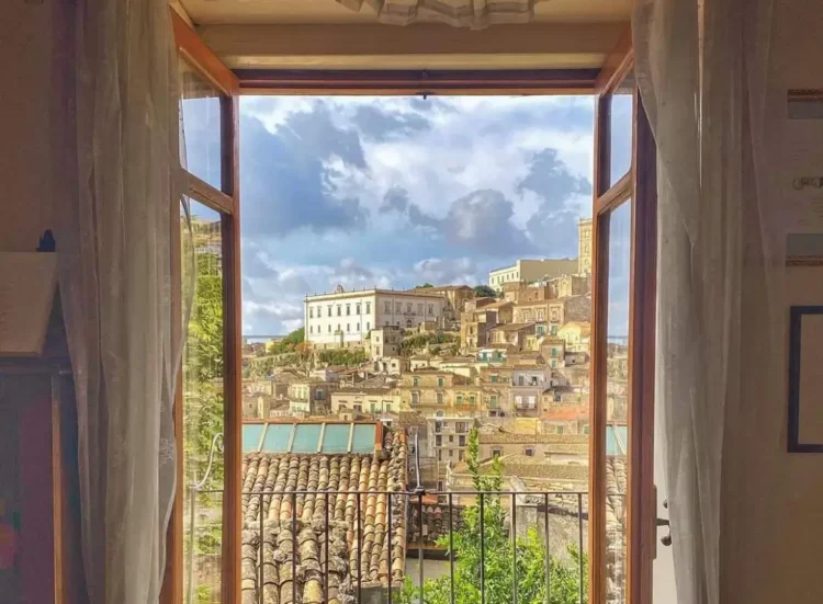 Vista dal centro storico di Modica attraverso una finestra aperta: tetti in cotto, un palazzo bianco e un campanile.