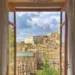 Vista dal centro storico di Modica attraverso una finestra aperta: tetti in cotto, un palazzo bianco e un campanile.